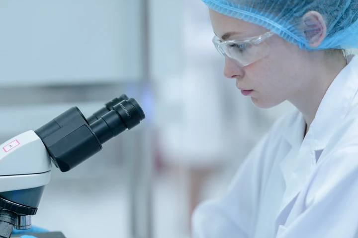Tecnica di laboratorio con occhiali protettivi e cuffia che osserva al microscopio